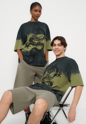 To modeller iført matchende grønne og svarte t-skjorter med dragegrafikk; den ene står med hendene i lommene, den andre sitter på en metallstol.