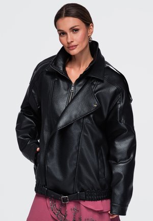 Femme aux cheveux bruns portant une veste en cuir noire oversize sur une jupe rose avec des détails en sequins, posant devant un fond gris clair uni.
