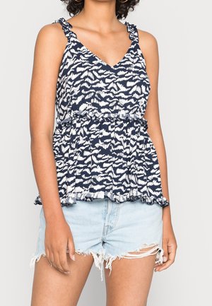 Mujer con un top sin mangas de patrón azul marino y blanco con detalles de volantes y shorts vaqueros azules claros desgastados.