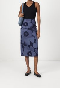 Žena v černém bezrukávovém topu, modré květinové midi sukni, černých plochých botách a modré kabelce stojící na bílém pozadí.