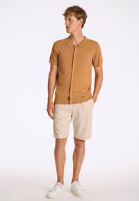 Rinnakkaisella kauluksella varustettu lyhythihainen, sinapinkeltainen neulepolo, jossa on nappilukko, yhdistettynä beigeihin shortseihin ja valkoisiin lenkkareihin. Yksinkertainen tausta.