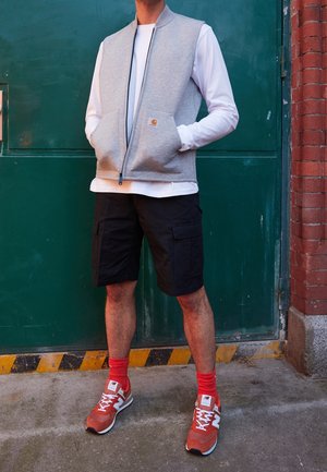 Osoba ubrana w jasnoszary bezrękawnik na zamek błyskawiczny, na białej koszuli z długim rękawem, czarne szorty cargo, czerwone skarpetki i pomarańczowe sneakersy New Balance, stojąca na zewnątrz.