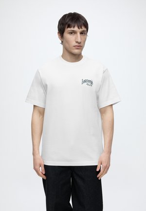 Junger Mann trägt ein weißes kurzärmeliges T-Shirt mit dem "Lacoste Paris"-Logo auf der Brust und schwarze Hose, steht vor einem schlichten Hintergrund.