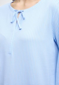 Blouse bleu clair avec des rayures verticales, un col rond, un détail noué au col et un tissu doux et texturé.