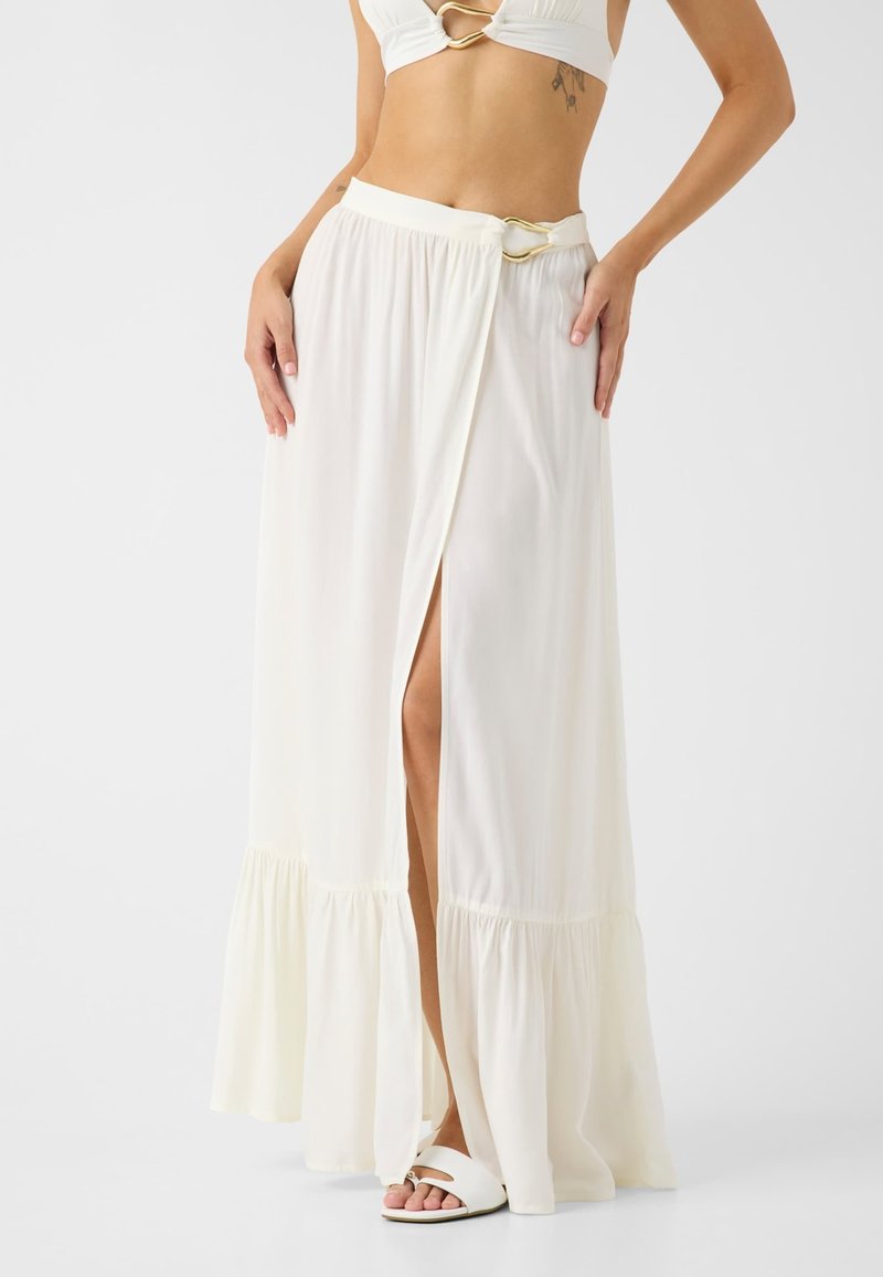 Femme portant une jupe longue blanche taille haute avec une fente frontale et une boucle dorée, associée à des sandales blanches et un top court.
