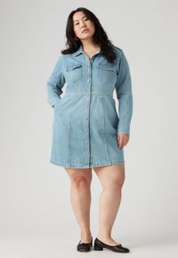 Ανοιχτό μπλε τζιν φόρεμα με κουμπιά μπροστά, μακριά μανίκια και δύο τσέπες στο στήθος. Διαθέτει εφαρμοστό μέσο και φαρδύ στρίφωμα.