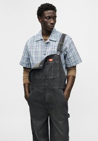 Νέος άνδρας που φοράει μαύρη τζιν φόρμα με ένα λουρί ξεκούμπωτο, μπλε πουκάμισο με κοντά μανίκια σε καρό σχέδιο και καφέ μπλουζάκι μακρυμάνικο από μέσα, με τα χέρια στις τσέπες.