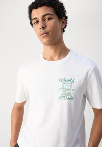 Bílý bavlněný tričko s krátkými rukávy, s zelenou grafickou potiskem budovy a textem. Design s kulatým výstřihem, hladká textura.