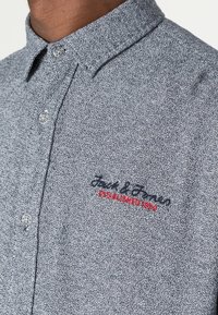 Košile s krátkým rukávem na knoflíky z šedé texturované látky. Má límec, dva knoflíky a vyšívané logo v tmavě modré a červené barvě.