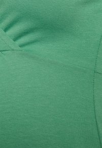 Tecido verde com uma textura suave; apresenta um estiramento subtil e costuras que criam um design ajustado. Cor uniforme sem padrões ou acentos visíveis.