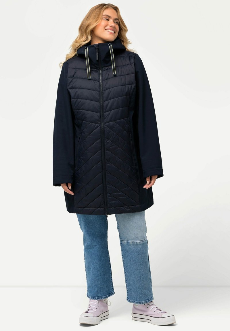 Cappotto lungo navy trapuntato con colletto alto, dettagli con cordino, due tasche laterali con zip e maniche ampie. Indossato sopra un denim chiaro e sneakers.