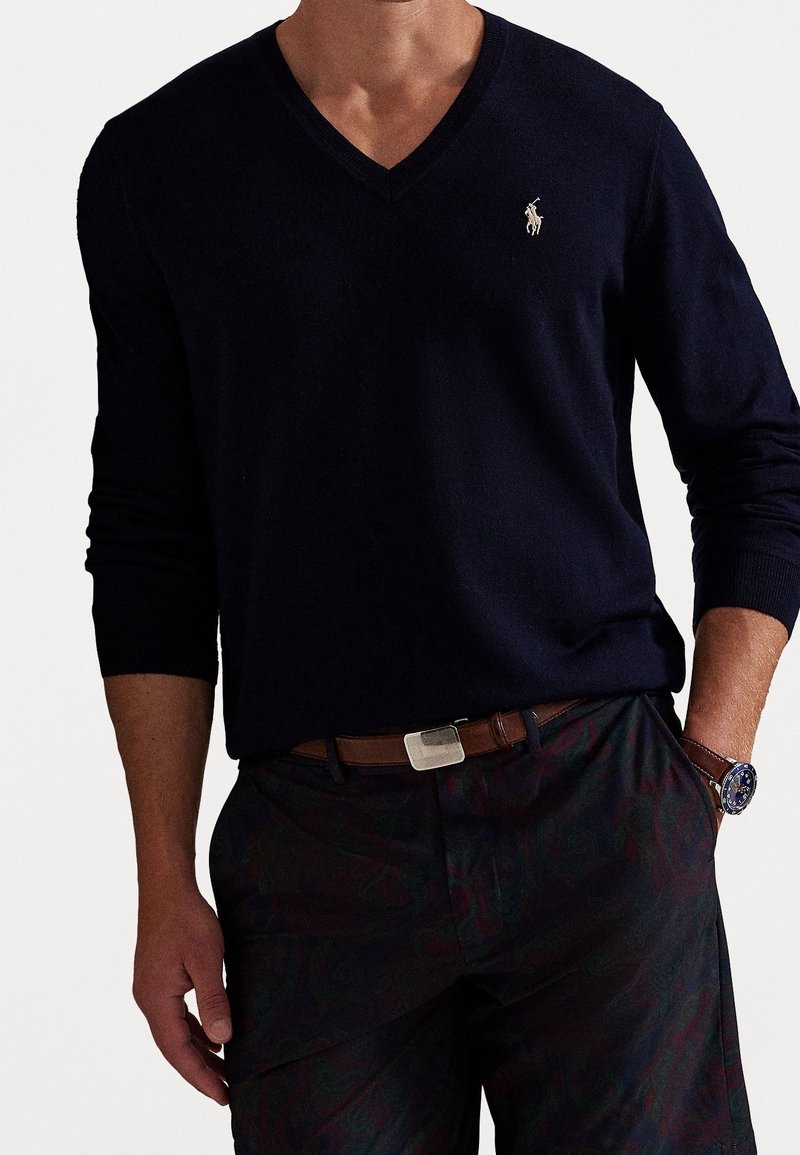 Homme portant un pull à col en V bleu marine foncé avec un petit logo Polo, un pantalon sombre à motifs, une ceinture marron et une montre-bracelet, la main dans la poche.