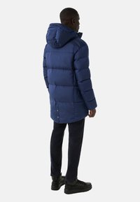 Mørkeblå dunjakke med hætte, der har et quiltet design, lang pasform og trykknaplukninger bagpå. Bæres over mørke bukser og støvler.