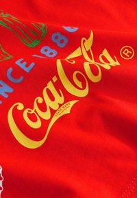 Czerwona tkanina z żółtym logo „Coca-Cola”, zieloną ilustracją butelki oraz niebieskim napisem „Since 1886” na gładkiej, tkanej fakturze.