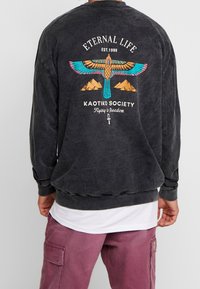 Felpa nera con una grafica di un'aquila in turchese e arancione. Presenta il testo: "VITA ETERNA", "KAOTIKO SOCIETY" e motivi di montagne.