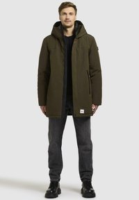 Olive green parka jacket with a hood, featuring a front zipper, snap closure, and two side pockets. Worn over a black shirt and dark jeans.