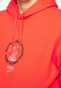 Röd hoodie av mjuk bomull, med en framsida som har en grafik med text och en tecknad figur. Inkluderar en huva med dragsko och en känguruficka.
