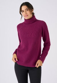 Suéter ajustado de cuello alto en magenta oscuro, con patrones texturizados en el canesú y puños acanalados, combinado con jeans negros.