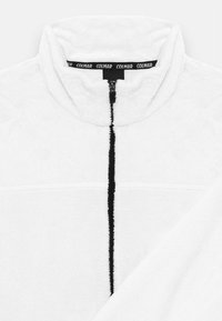 Biely fleecový zipsový cardigan s vysokým golierom, čiernym zipsom a horizontálnymi švovými detailmi cez hruď. Mäkká textúra, minimalistický dizajn.