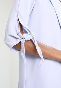 Blouse lavande claire avec manches nouées. Tissu doux, coupe décontractée, avec des ouvertures de manches accentuées et des détails subtils dans le design.