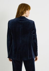 Blazer de terciopelo azul marino con cuello con muesca, corte entallado y puños con botones. La parte trasera tiene una sola abertura y una textura lisa.