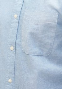 Camisa azul clara de algodão com botões, apresenta um bolso aplicável no lado esquerdo e botões de plástico branco. Textura suave, design casual.