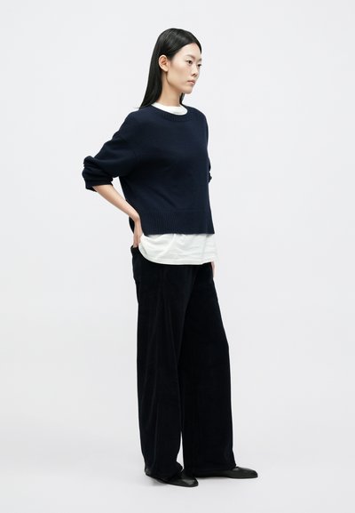 Ναυτικό πλεκτό πουλόβερ με ριμπ τελείωμα, φορεμένο πάνω από λευκό πουκάμισο, συνδυασμένο με μαύρο παντελόνι με φαρδιά μπατζάκια και μαύρες μπαλαρίνες. Απλός, μοντέρνος σχεδιασμός.