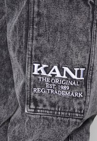 Tela de mezclilla negra con textura desgastada. Presenta un logo bordado en blanco con los detalles "KANI THE ORIGINAL EST 1989" y "REG. TRADEMARK."