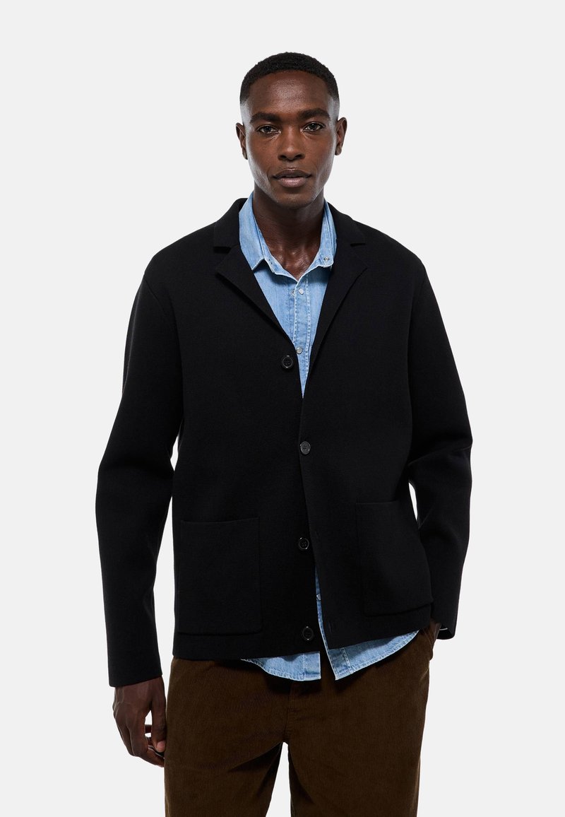 Veste en laine noire avec un col à revers, fermeture à bouton sur le devant et deux poches avant, portée sur une chemise en denim bleu clair.