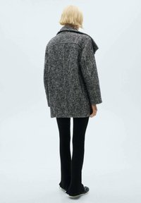 Manteau en tweed gris et noir à coupe décontractée, doté d'un col à revers et d'une grande fente au dos. Porté avec un pantalon noir côtelé et ajusté.