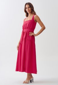 Fuchsia maxi dress with square neckline, elastic waist, and pleated detail. Paired with white strappy heeled sandals. Smooth fabric texture.