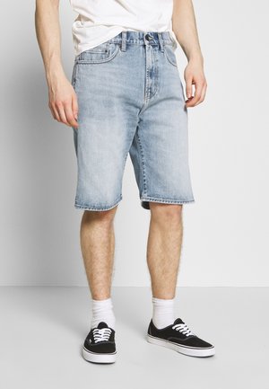 Person trägt hellblaue Jeansshorts, weiße Socken und schwarze Low-Top-Sneaker und steht vor einem schlichten Hintergrund.