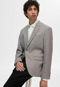 Ljusgrå blazer i texturerad tyg med slagkrage, enkel knappstängning och två framfickor. Bärs över en vit skjorta.