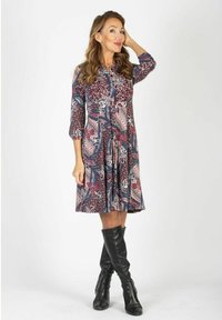 Kobieta z długimi falującymi włosami, ubrana w wzorzystą sukienkę do kolan oraz czarne kozaki za kolano, stojąca z ręką dotykającą włosów.