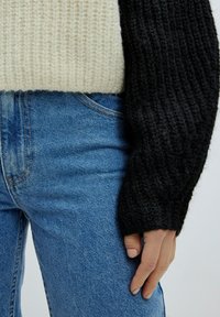 Gebreide trui met een beige lichaam en zwarte geribbelde mouwen, gecombineerd met blauwe denim jeans. Close-up van texturen en kleuren zichtbaar.