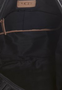Intérieur en tissu noir d'un sac avec une étiquette logo UGG. Présente une texture lisse et un design minimaliste. Détail de poche visible.