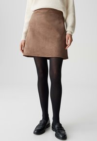 Brauner Wildleder-A-Linien-Rock mit glatter Textur und schlichtem Saum, kombiniert mit schwarzen Strumpfhosen und Slippern, getragen mit einem leichten Strickpullover.