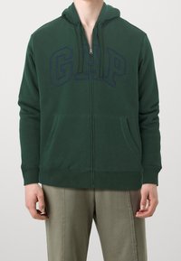 Sudadera verde con cremallera fabricada en mezcla de algodón, presenta un gran logo de "GAP" en color marino en el frente, bolsillos frontales y capucha con cordones.