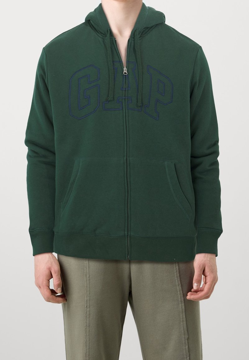 Felpa verde con zip realizzata in misto cotone, caratterizzata da un grande logo "GAP" in blu navy sul davanti, tasche frontali e cappuccio con cordino.