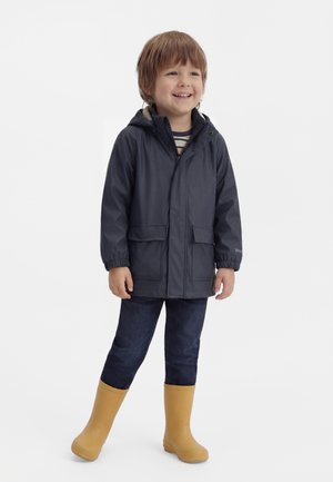 Uśmiechnięty młody chłopiec w granatowej kurtce przeciwdeszczowej, ciemnych dżinsach i żółtych kaloszach, stojący na jednolitym białym tle.