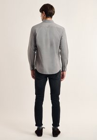 Camisa gris de manga larga con puños de botón, que presenta un cuello clásico y un yugo trasero, combinada con jeans negros ajustados y zapatos marrones.