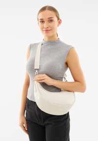 Borsa hobo in materiale liscio di colore crema con un design curvo. La tracolla beige regolabile presenta un testo ed è indossata da un modello con una maglietta grigia.