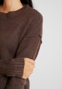 Hnědý pletený svetr s žebrovan�ými manžetami, s kulatým výstřihem a měkkou, texturovanou tkaninou. Detail ruky s překříženými pažemi.