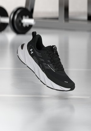 Must-valge Under Armour jooksujalats ujub peegeldava jõusaalipõranda kohal, kus taustal on hägused raskused.