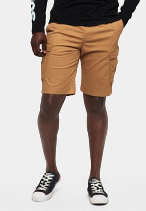 Mann trägt beige Cargoshorts, schwarzes Langarmshirt und schwarze Sneakers mit weißen Sohlen und Schnürsenkeln, steht vor weißem Hintergrund.
