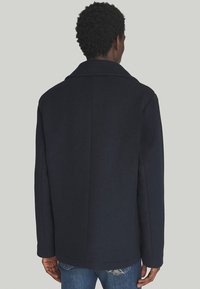 Cappotto di lana blu navy con colletto a rever, caratterizzato da una linea dritta e senza chiusure visibili. Tessuto liscio, design di lunghezza media.