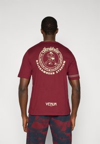 Μπορντό βαμβακερό μπλουζάκι με γραφική εκτύπωση και κείμενο στην πίσω πλευρά. Χρυσές λεπτομέρειες αναδεικνύουν το σχέδιο. Κοντά μανίκια και άνετη εφαρμογή.