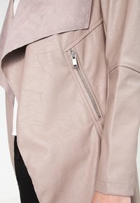 Manga de chaqueta de cuero color topo con bolsillo de cremallera plateada visible y detalle de cuello de ante drapeado, llevada sobre ropa blanca y negra.