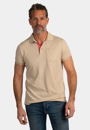 Man met kort grijs haar en baard, gekleed in een beige poloshirt met roze kraagdetail en een blauwe spijkerbroek, staand tegen een effen achtergrond.