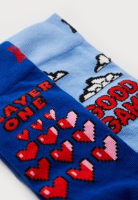 Chaussettes en coton bleues et bleu foncé avec des motifs de cœurs pixelisés et du texte : "JOUEUR UN" en rouge et "BON JEU" en rouge avec un contour noir.